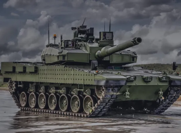 Yeşil kamuflajlı bir ana muharebe tankı (Altay), bulutlu gökyüzü altında sahada duruyor, namlusu ileriye dönük