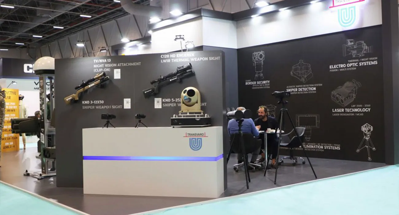 Fuar standında duvara monte edilmiş KND 3-12x50 ve KND 5-25x56 keskin nişancı dürbünleri ile C120HD ENGEREK termal silah dürbünü; önde iki kişi bir masada otururken arkada ürün kategori şeması görünüyor