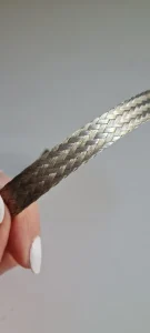 Bir elin iki parmağı arasında tuttuğu, dikey duran yassı örgülü metal şerit, gri arka plan