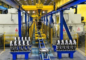 Sarı portal robotun altında, mavi rayların iki yanında sıralanmış parlak işlenmiş demiryolu tekerlekleri bulunan teker işleme hattı